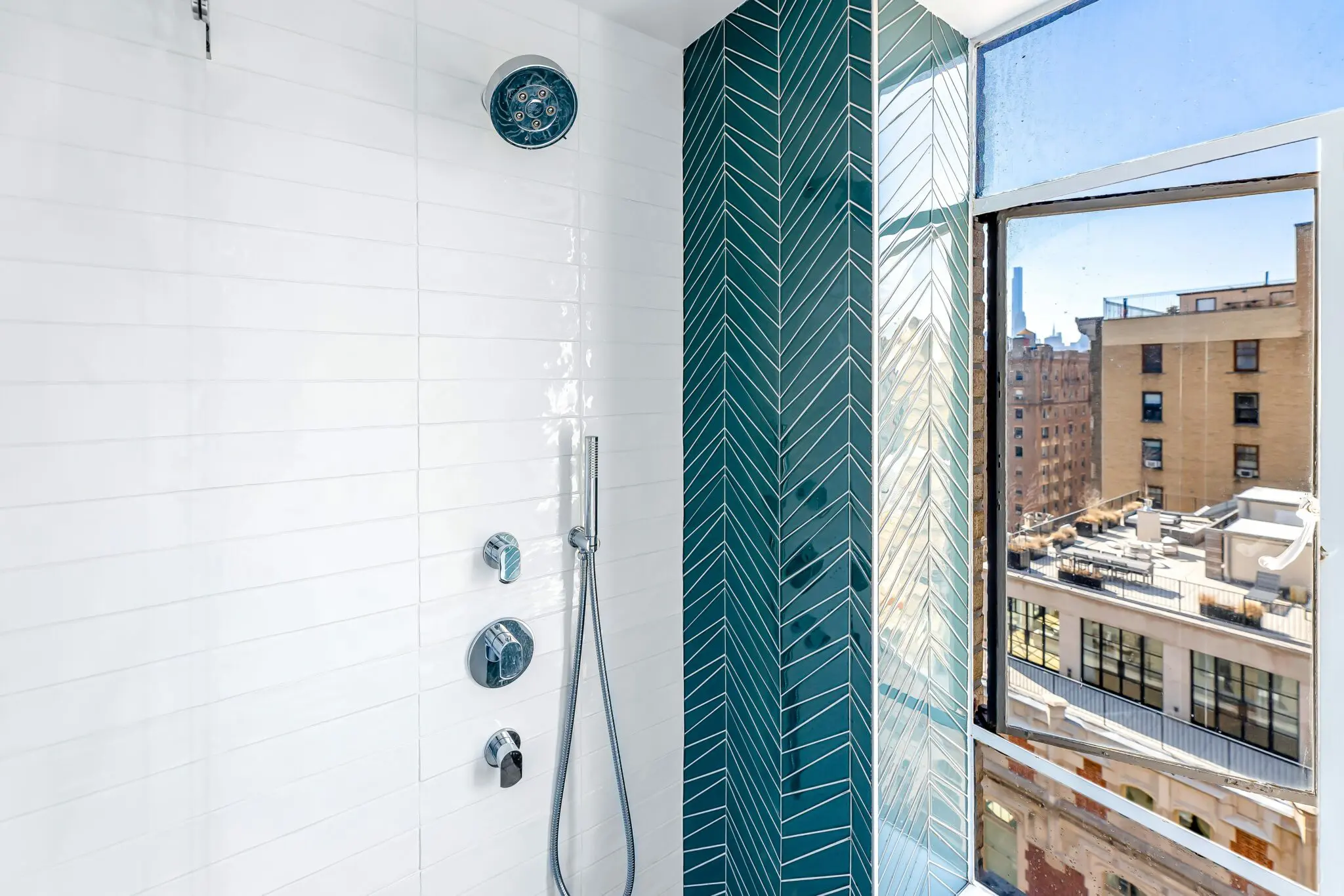 Modern shower with white tiles and a vertical teal herringbone accent wall. Chrome shower fixtures include a round showerhead and handheld spray. A large window offers a view of city buildings under a clear blue sky.