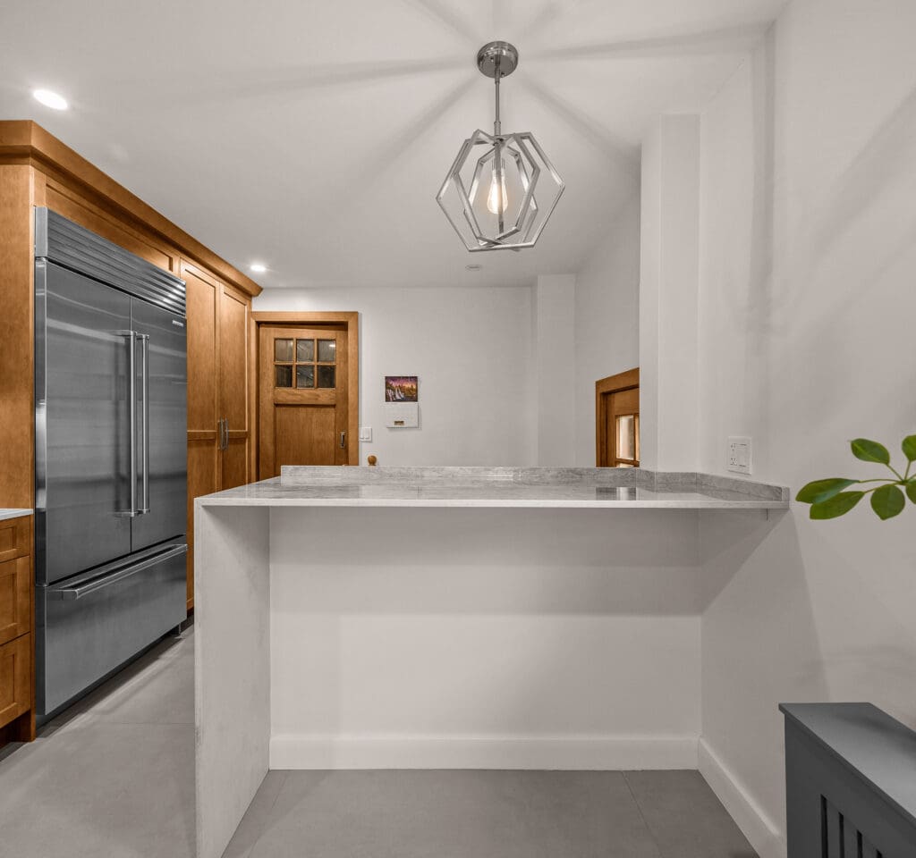 A modern kitchen with a large stainless steel refrigerator, wooden cabinets, and a marble-topped island. A geometric pendant light hangs from the ceiling. A small plant is in the corner, and a wooden door is visible in the background.