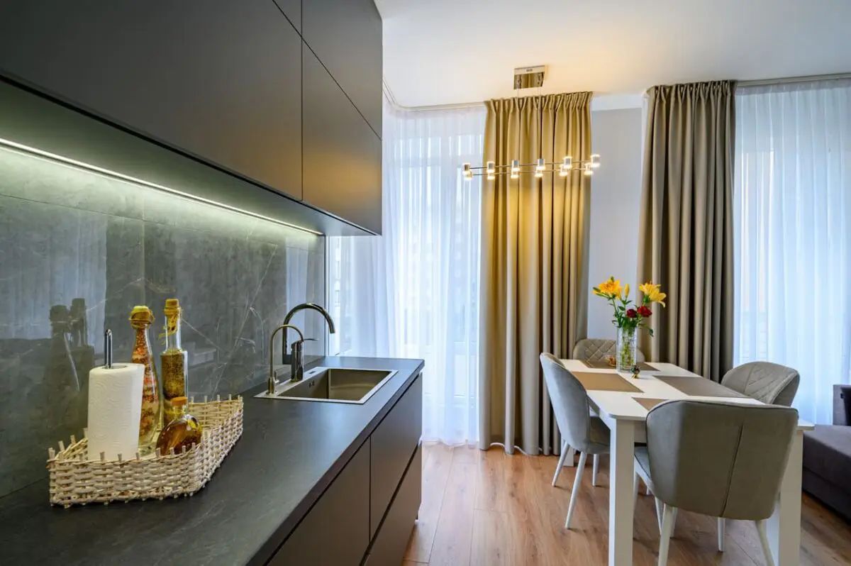 Contemporary kitchen featuring matte-finished black cabinets paired with a sleek gray stone backsplash and countertop. Under-cabinet lighting enhances the modern aesthetic, while the adjacent dining area with neutral tones and soft drapery creates a cozy and inviting atmosphere