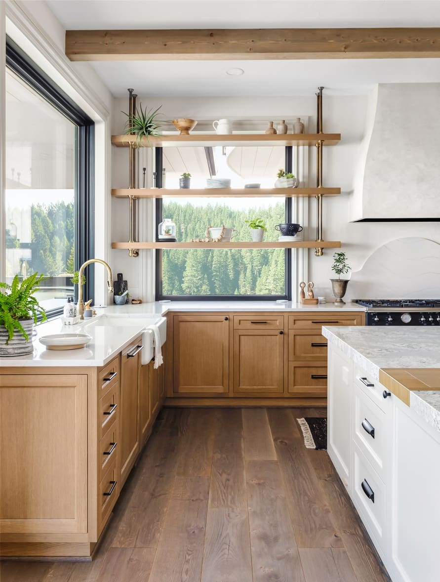 A modern kitchen showcases excellent kitchen cabinet choices with light wood cabinets, a farmhouse sink, white countertops, and open shelves adorned with plants and dishes. Large windows reveal a forest view, while the wooden floor complements a stove featuring a marble backsplash and white hood.