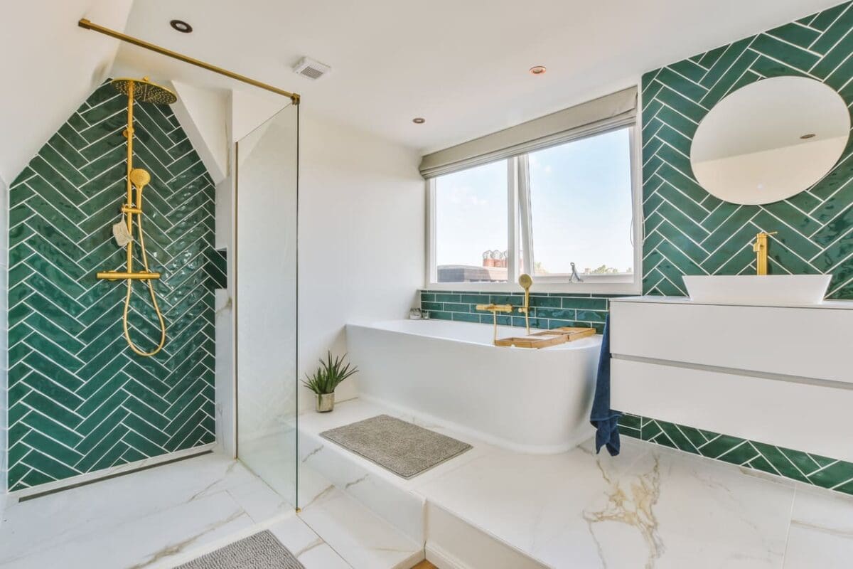 Bathroom featuring green chevron-patterned tile layout on walls, a gold rain shower head, a freestanding white tub with matching gold fixtures, and a modern white floating vanity with a circular mirror. Perfect inspiration for a chevron tile layout