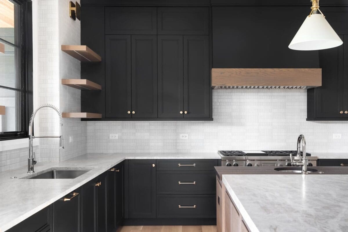 Luxurious kitchen featuring inset cabinets in a sleek black finish with gold hardware, paired with a white marble countertop and backsplash. Open wooden shelves and a custom wood range hood add warmth, while a polished gooseneck faucet and stainless steel appliances complete the elegant, tailored design