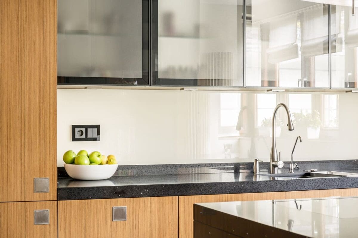 A modern kitchen boasts a sleek Glass-Front Cabinets design featuring wooden cabinets that highlight the diverse kitchen cabinet choices. A black countertop and stainless steel faucet complement the decor, while a white bowl of green apples and natural light enhance the minimalist charm.