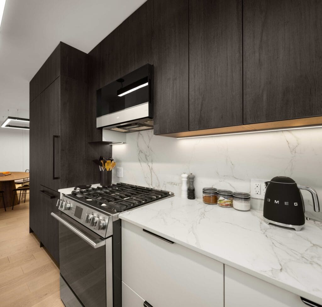 Modern kitchen renovated by NYKB with black cabinets and a marble countertop. Features a stainless steel gas stove, kettle, and spice jars. Well-lit with recessed lighting under cabinets. A dining area with a wooden table is visible in the background.
