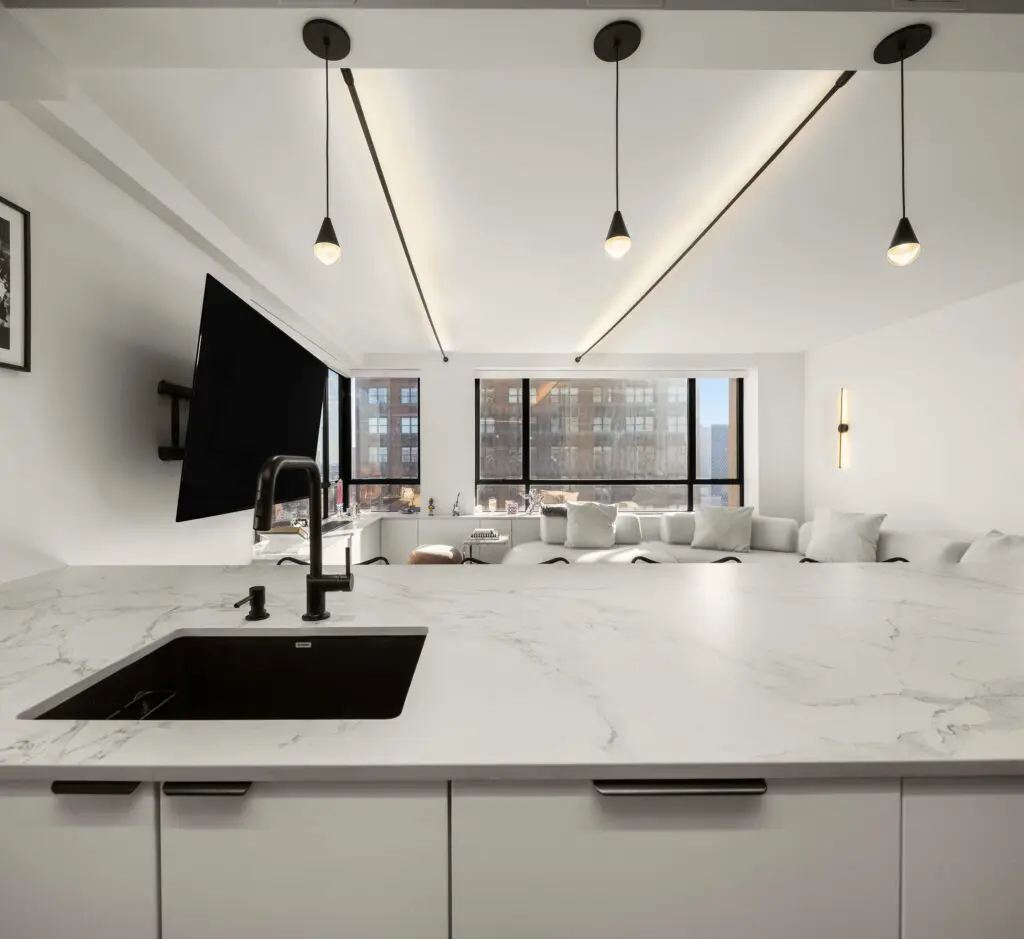 Modern kitchen with a marble countertop and black sink. Minimalist hanging lights above the island. In the background, theres a living area with a wall-mounted TV, white sofa, and large windows with a city view.