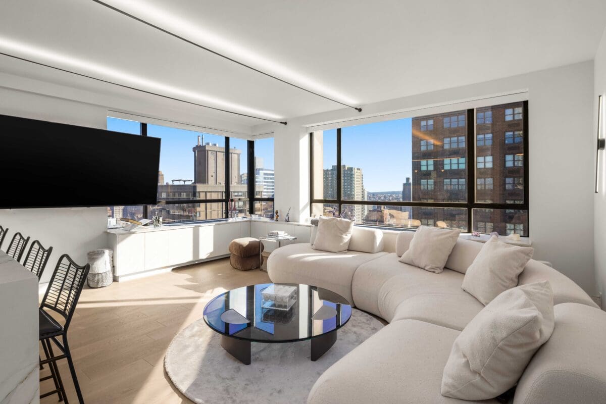 Spacious modern living room, the result of a full home renovation, boasts large windows showcasing a city view. Features include a curved white sofa, glass coffee table, wall-mounted TV, and bar stools along a sleek counter. Natural light floods in, highlighting the contemporary decor.