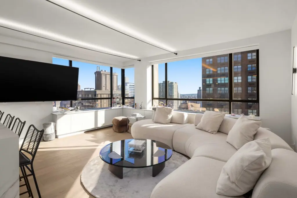 A modern living room with large windows, a curved beige sofa, a circular glass coffee table, and a wall-mounted TV. The room offers a panoramic view of urban buildings and a clear blue sky.