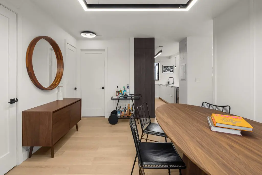 A modern dining area with a wooden oval table, four black wire chairs, and a sideboard with a round mirror above it. A small bar cart is placed near the door. The room has light wooden flooring and white walls with contemporary decor.