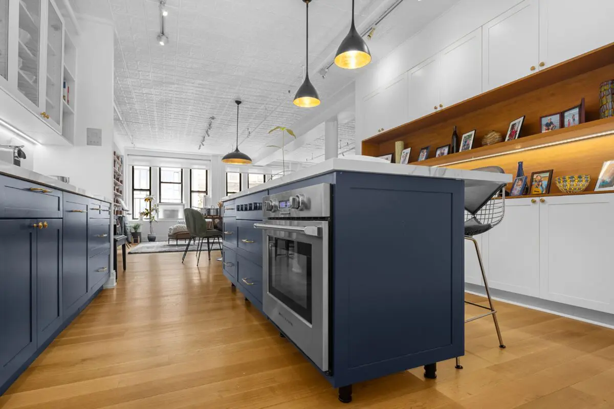 Modern kitchen with blue cabinetry and a central island featuring an oven. Pendant lights hang from a white, textured ceiling. The room extends into a living area with windows and shelves displaying pictures and decor. Hardwood floors throughout.