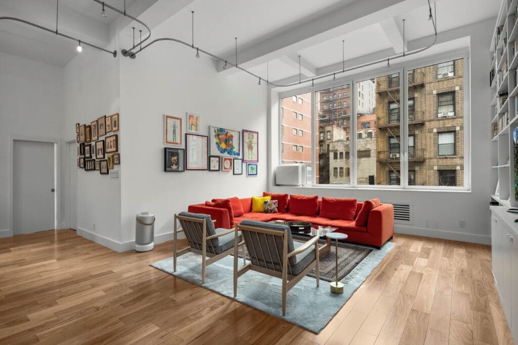 Modern living room created by nykb design and renovation company, featuring a red sectional sofa and two gray armchairs on a light blue rug. The room features wooden floors, large windows showing city buildings, and a collection of artwork on the walls. Ceiling lights hang above.