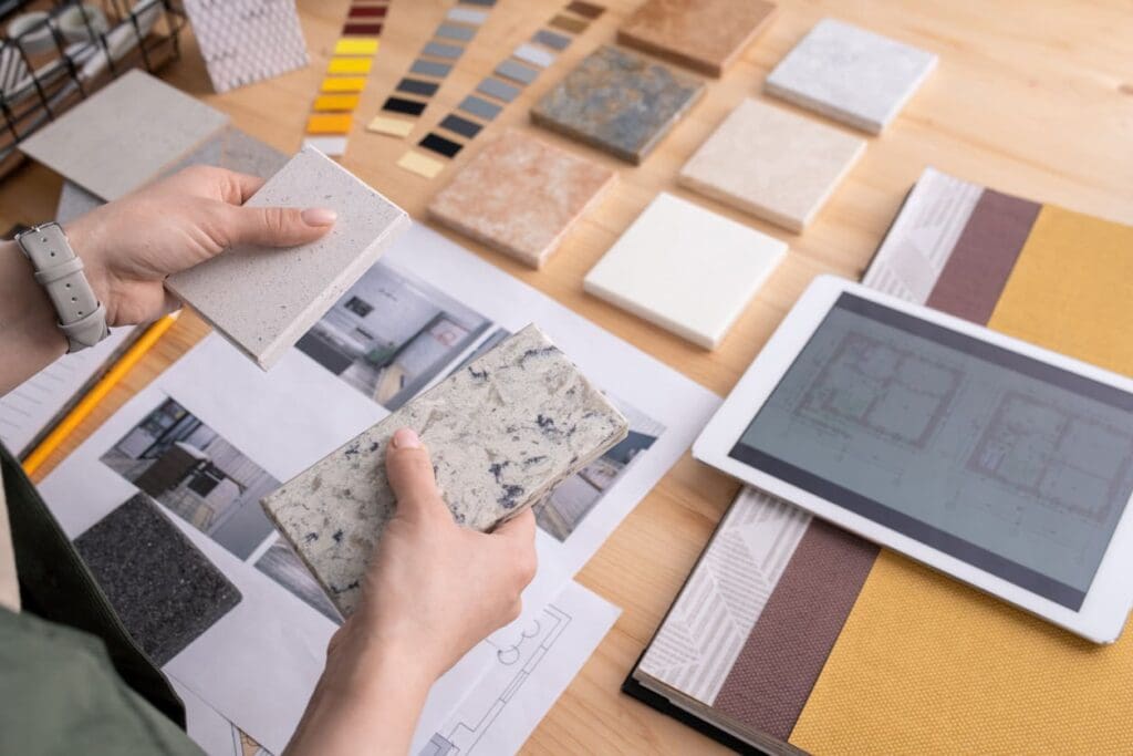 A person holds two stone samples over a table with more samples, a tablet displaying building plans, and a color swatch. The table also has design papers and fabric samples, indicating a design or renovation project.