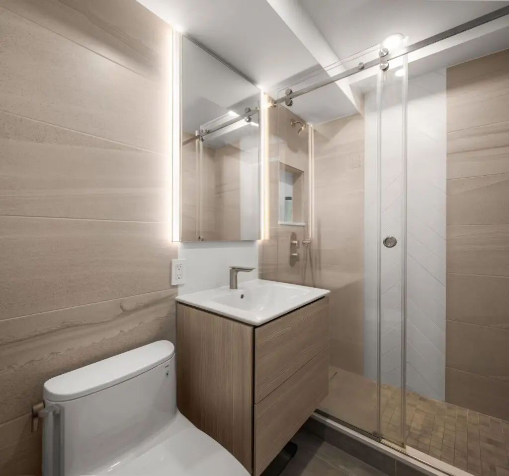Modern bathroom featuring a toilet and a wooden vanity with a sink. A large mirror with integrated lighting hangs above. On the right, a glass-enclosed shower with a sliding door, white tiles, and a tiled floor is visible.