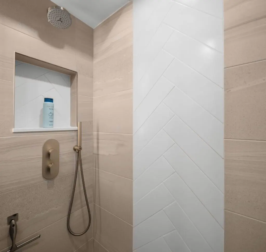 Modern shower with beige and white tile design. Wall niche holds a bottle of hair care product. Ceiling-mounted showerhead and handheld attachment with a sleek bronze faucet.