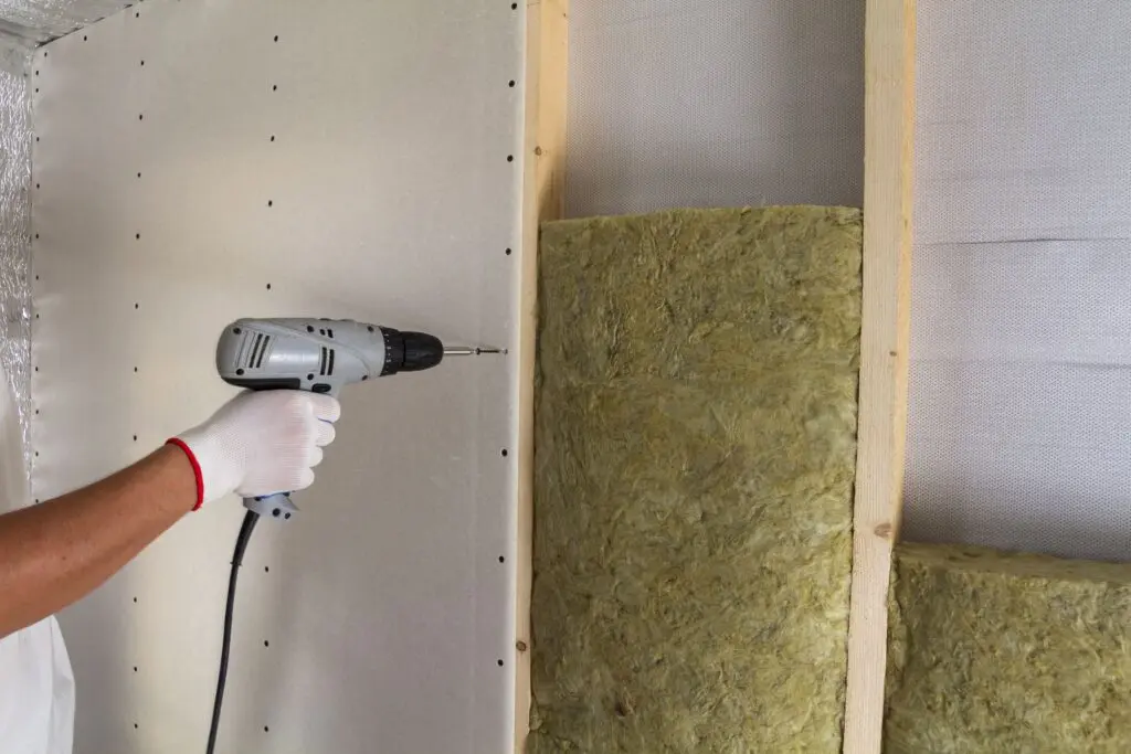 Person using a power drill to attach drywall to wooden studs, with yellow insulation visible between the studs. The person is wearing a white glove on their hand, holding the drill against the drywall.