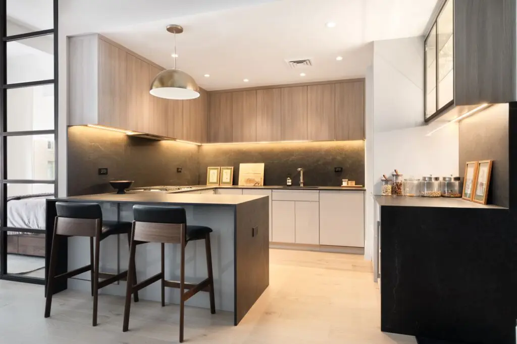 Modern kitchen with light wood cabinets, black countertops, and an island with two dark wooden barstools. Pendant light hangs above the island; framed art and jars decorate the counters.