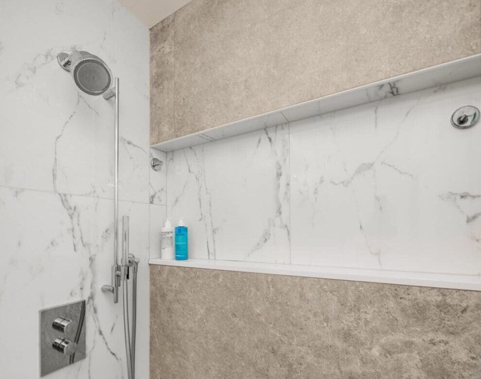 Modern shower with white marble and beige stone walls, a rainfall showerhead, handheld shower, built-in shelf holding two bottles, and a silver control panel. The floor features small square white tiles. Built in shower niche