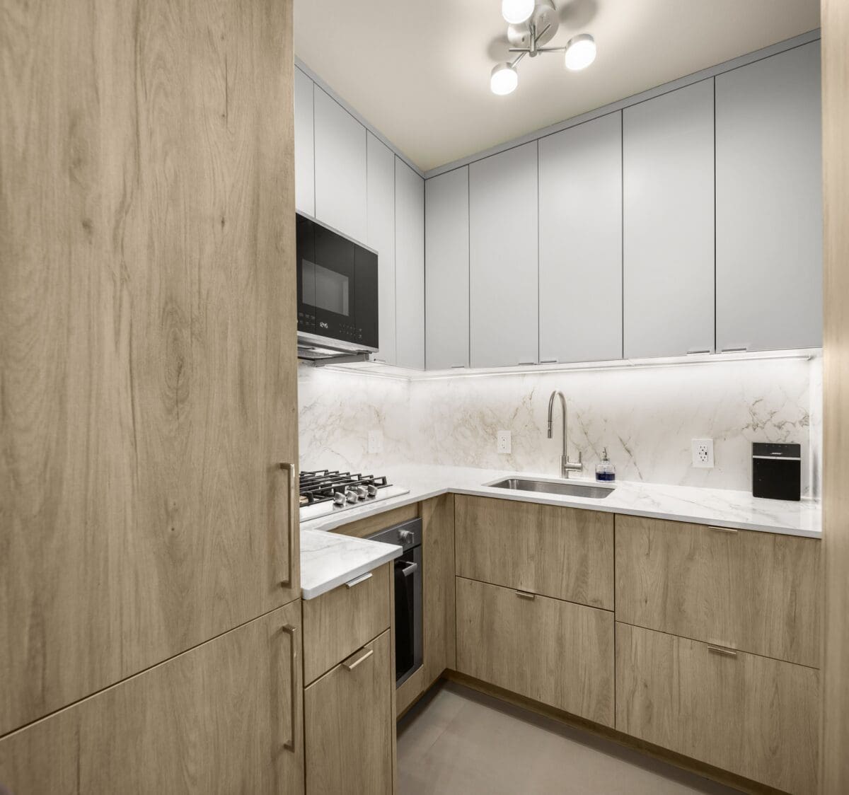 Modern kitchen with light wood cabinets, gray upper cabinets, marble countertops, built-in gas stove, microwave, and under-cabinet lighting. A single-basin sink is set beneath a minimalist faucet.