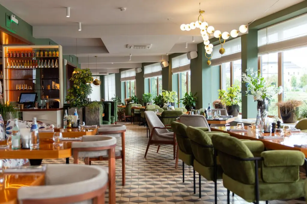 A stylish restaurant interior, crafted through expert Commercial Renovation in NYC, features green upholstered chairs, wooden tables set for dining, large windows, potted plants, and modern lighting for a bright and inviting atmosphere.