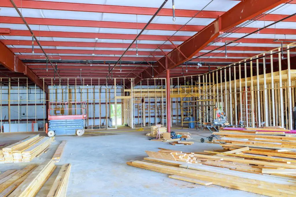 Interior of a building under construction in NYC, showcasing exposed metal beams and wooden framing. Stacks of lumber and equipment scattered on the concrete floor highlight an active commercial renovation in progress.