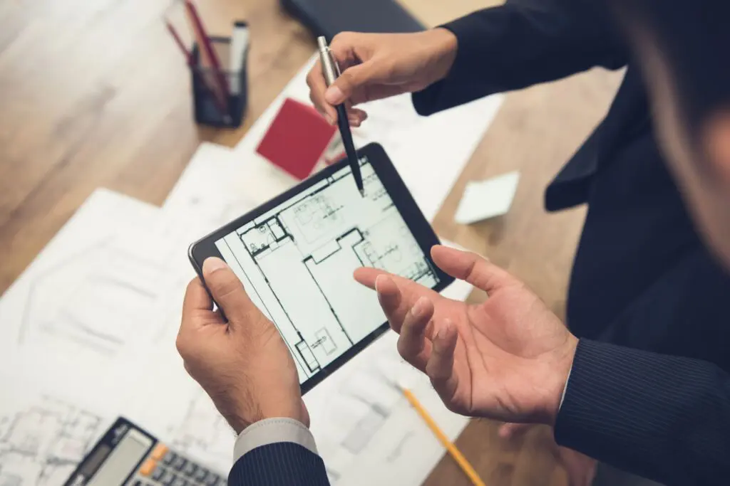 Two people in business attire discuss a commercial renovation in NYC, reviewing a building floor plan on a tablet. Architectural drawings, a calculator, and office supplies are spread across the wooden table.
