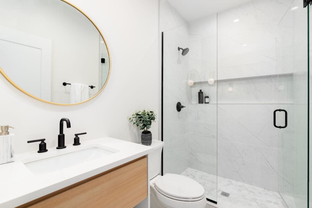 Modern bathroom with a round mirror above a white sink, black fixtures, a light wood vanity, a small potted plant, white walls, and a glass walk-in shower and built in shower shelves with black hardware.