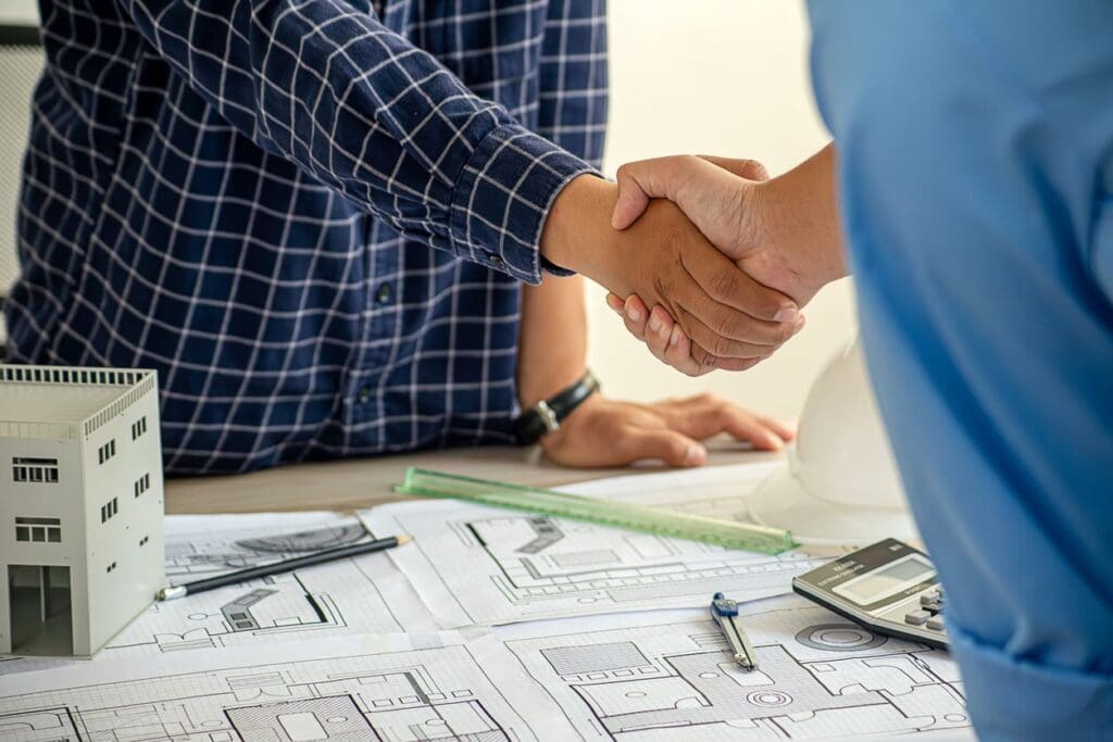 Two people shake hands over a desk with architectural blueprints, a model building, a hard hat, a ruler, and a calculator, suggesting a business agreement or partnership in architecture or construction.