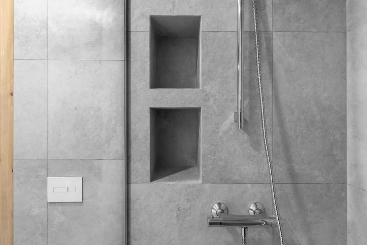 A modern shower with light gray tiled walls, two rectangular recessed shelves, a chrome showerhead and hose, and a minimalistic white flush plate on the left.