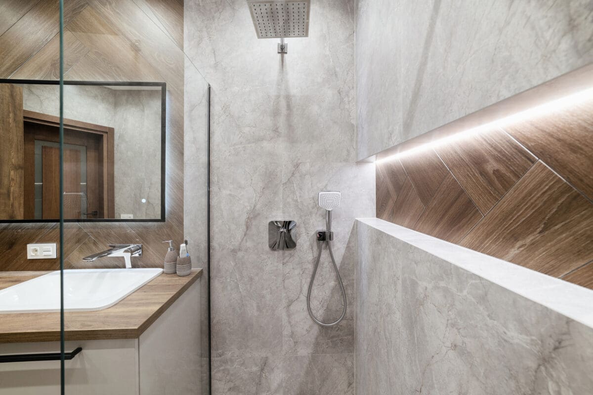 Modern bathroom with light gray marble walls, wood accents, a wall-mounted shower head, handheld shower, shower niche with LED lighting, and a white sink on a wooden countertop with soap dispensers.