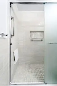 A modern walk-in shower with light beige tiles, a frosted glass door, built-in wall shelf, grab bar, and foldable seat. This minimalist setup offers accessible design for NYC apartments while maintaining style and functionality.