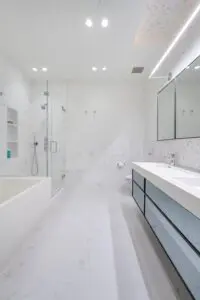 Modern white bathroom with accessible design for NYC apartments, featuring a glass-enclosed shower, built-in bathtub, double sink vanity with drawers, and an illuminated mirror above. Marble floors and walls complete the minimal decor.