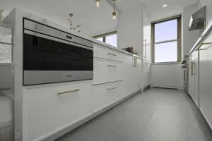 Modern kitchen with white cabinets, gold handles, stainless steel built-in oven, large gray floor tiles, and two windows letting in natural light. Contemporary light fixtures hang from the ceiling.