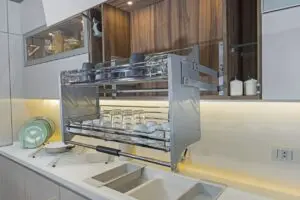 A modern kitchen features a pull-down dish rack above a sink, holding glasses and plates. Green plates and bowls are stacked on a drying rack beside the sink. The backsplash and cabinets are white.