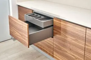 A modern kitchen cabinet with a wooden finish has an open drawer revealing a gray cutlery organizer tray inside. The countertop is light-colored, and the floor is light wood.