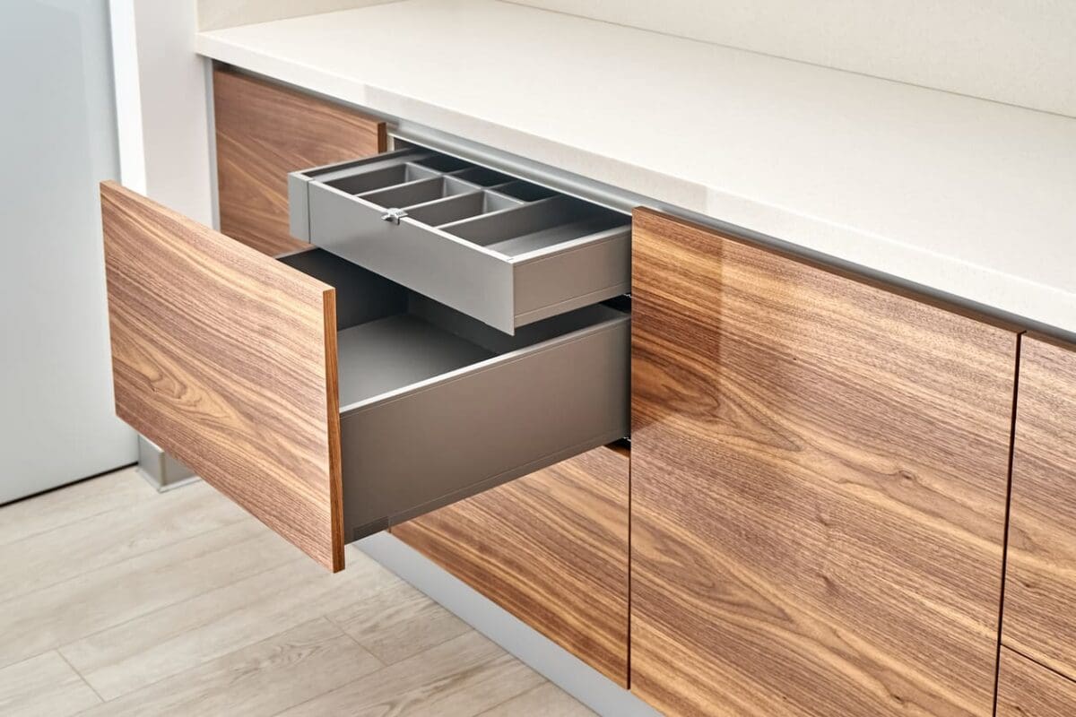 A modern kitchen cabinet with a wooden finish has an open drawer revealing a gray cutlery organizer tray inside. The countertop is light-colored, and the floor is light wood.