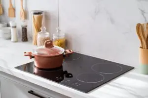 A modern kitchen with a black glass induction cooktop, a covered copper-colored pot on one burner, and jars of pasta, grains, and utensils arranged neatly on a marble countertop and backsplash.