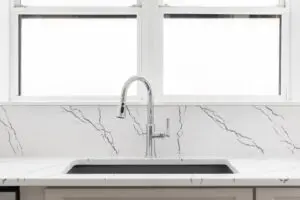 A modern kitchen sink with a curved chrome faucet set in a white marble countertop with gray veining, positioned in front of two large, frosted windows.