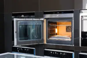 A modern built-in electric oven with its door open, showing empty wire racks and the interior light on. The oven is installed within a glossy black kitchen cabinet.