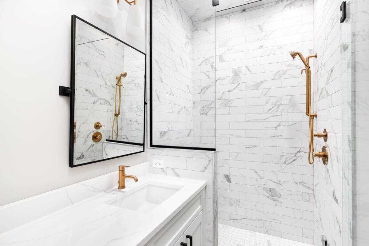 Modern bathroom with white marble tiles, a glass shower enclosure, gold fixtures including faucets and showerhead, a rectangular mirror, and a white vanity with double sinks.