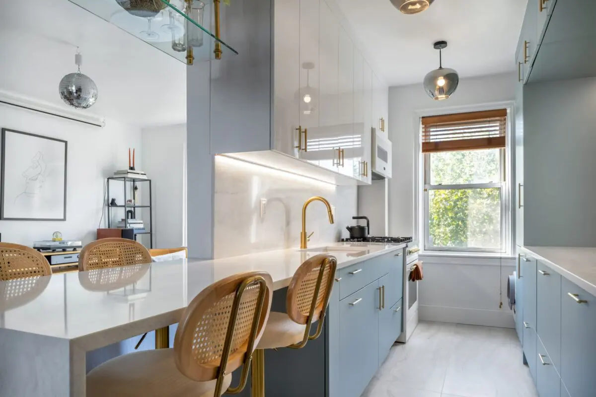 Modern kitchen with light blue cabinets, gold hardware, white countertops, and under-cabinet lighting. Three wooden barstools sit at an island. A window lets in natural light, with a dining area visible in the background.
