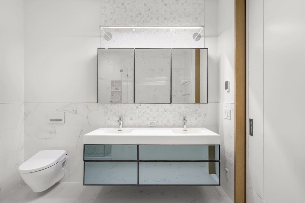 Modern bathroom with a floating vanity featuring two sinks, a large mirrored cabinet above, a wall-mounted toilet on the left, and white marble walls and floor for a clean, minimalist look.