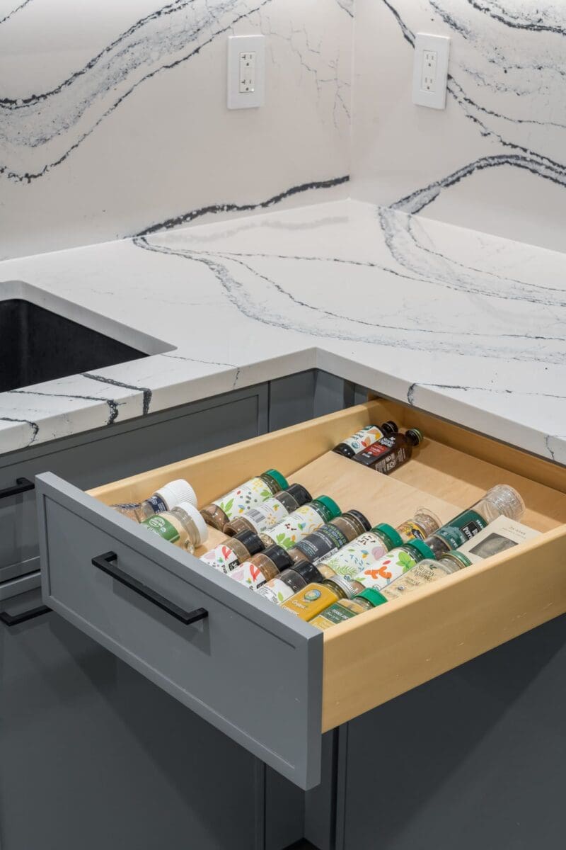 A kitchen drawer partially open in a gray cabinet reveals neatly organized spice jars. The countertop is white with dark, wavy veining, and there is a black sink and two electrical outlets visible.
