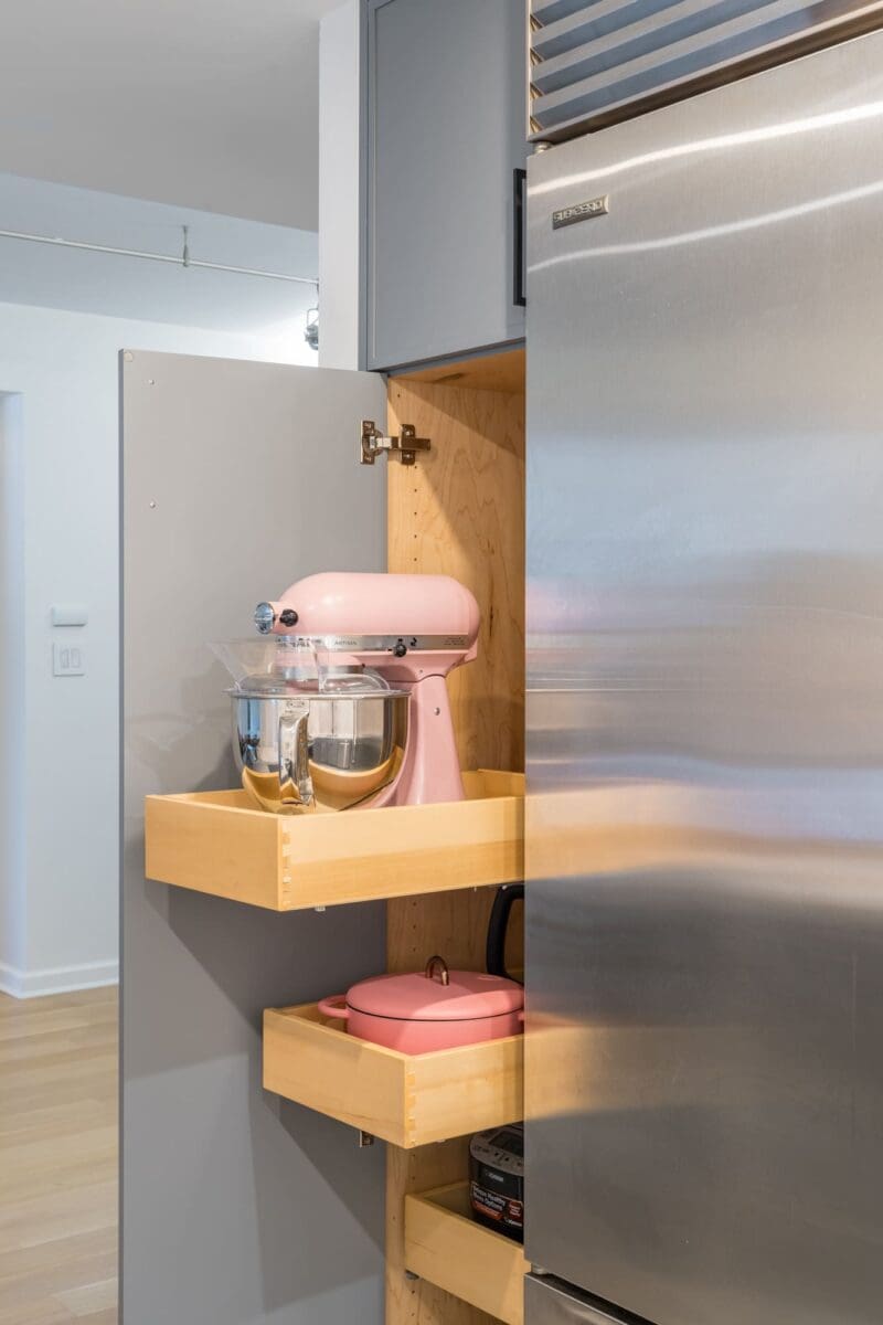 A cabinet with two pull-out wooden shelves holds a pink stand mixer and mixing bowl on the top shelf, and a pink dutch oven on the lower shelf, next to a stainless steel refrigerator.