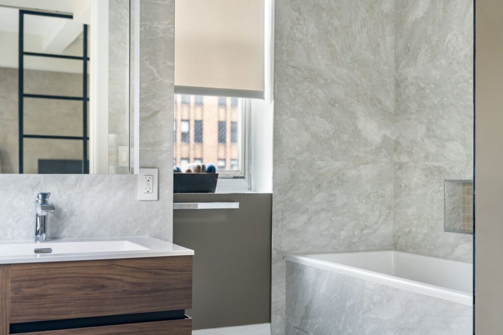 Modern bathroom with gray marble walls, a rectangular white bathtub, wood vanity with a sink and faucet, large mirror, and a window with a beige roller shade letting in natural light.