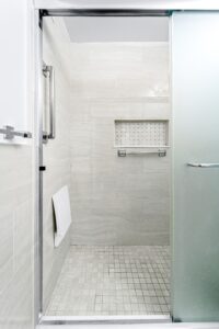 A modern walk-in shower with light beige tiles, a frosted glass door, built-in wall shelf, grab bar, and foldable seat. This minimalist setup offers accessible design for NYC apartments while maintaining style and functionality.