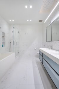 Modern white bathroom with accessible design for NYC apartments, featuring a glass-enclosed shower, built-in bathtub, double sink vanity with drawers, and an illuminated mirror above. Marble floors and walls complete the minimal decor.