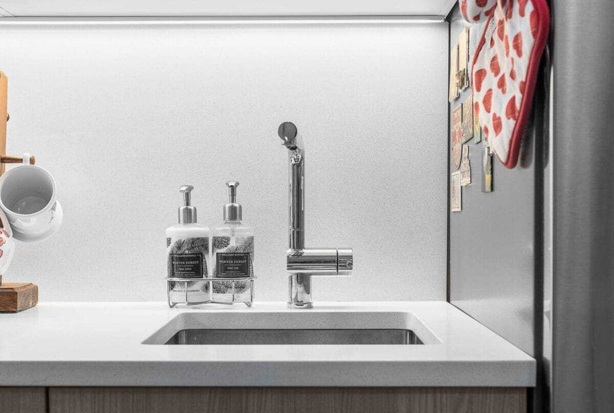 A modern kitchen sink with a sleek metallic faucet. Beside the sink are two soap dispensers with labels. A rack with mugs is partially visible on the left. On the right, theres a fridge with decorative magnets and oven mitts hanging.