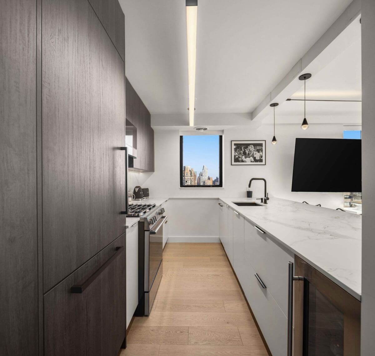 Modern kitchen with sleek dark wood cabinets and white countertops. Features a stainless steel stove, large window with a city view, pendant lights, and framed wall art. Light wood flooring and a built-in oven complete the contemporary look.