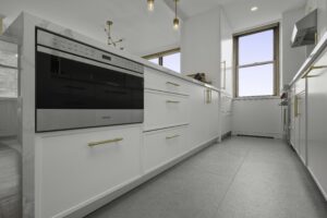 Modern kitchen with white cabinets, gold handles, stainless steel built-in oven, large gray floor tiles, and two windows letting in natural light. Contemporary light fixtures hang from the ceiling.
