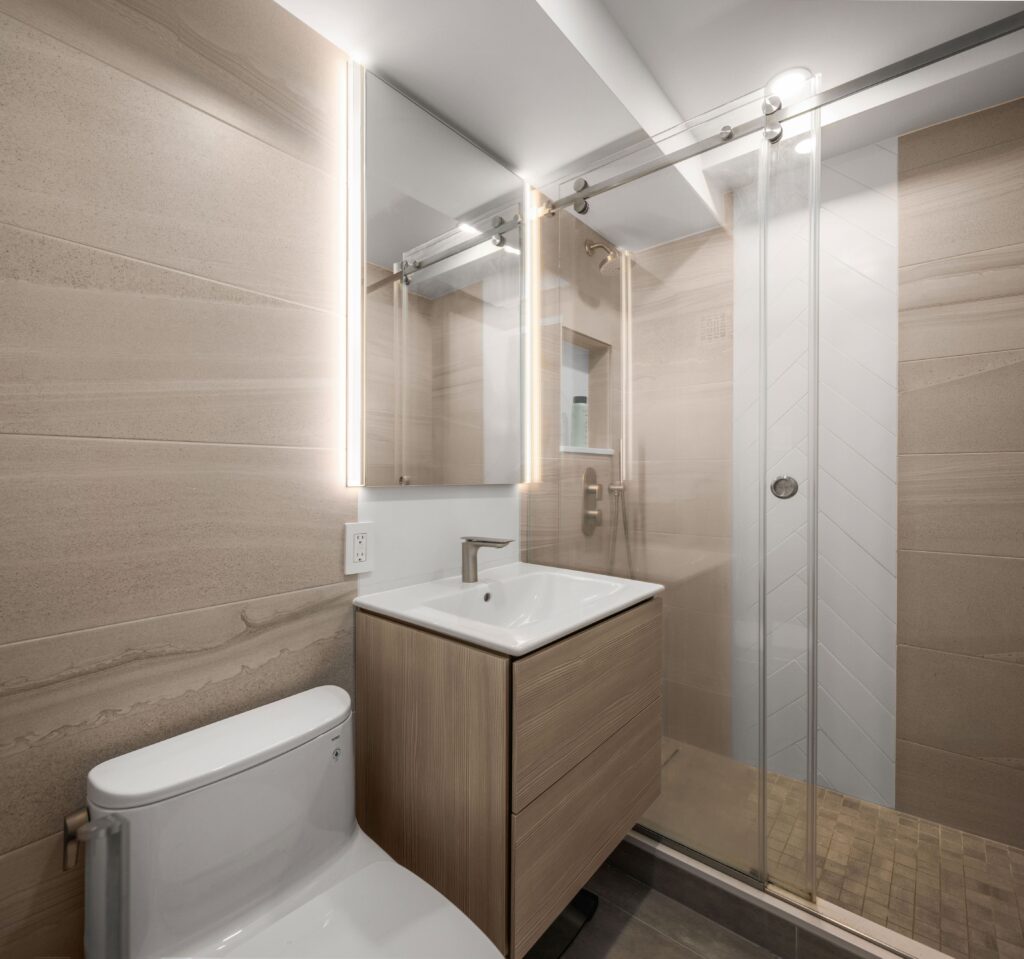 Modern bathroom featuring a toilet and a wooden vanity with a sink. A large mirror with integrated lighting hangs above. On the right, a glass-enclosed shower with a sliding door, white tiles, and a tiled floor is visible.