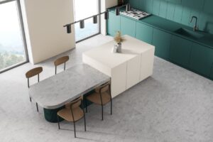 Modern kitchen with teal cabinets, a white island, and a gray countertop. Four wooden chairs are arranged around the island, and a vase with dried flowers sits on top. Large windows let in natural light.